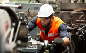 Man working on machine with safety hat, vest, glasses, and gloves on.